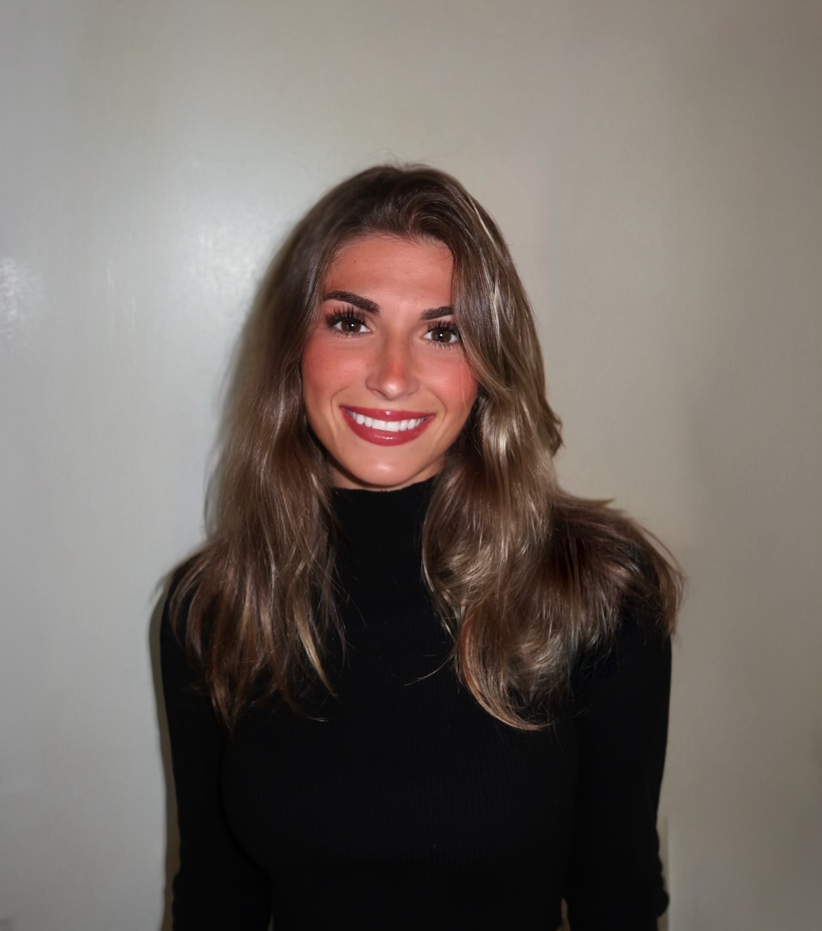 A smiling woman with wavy hair wearing a black top against a plain wall.