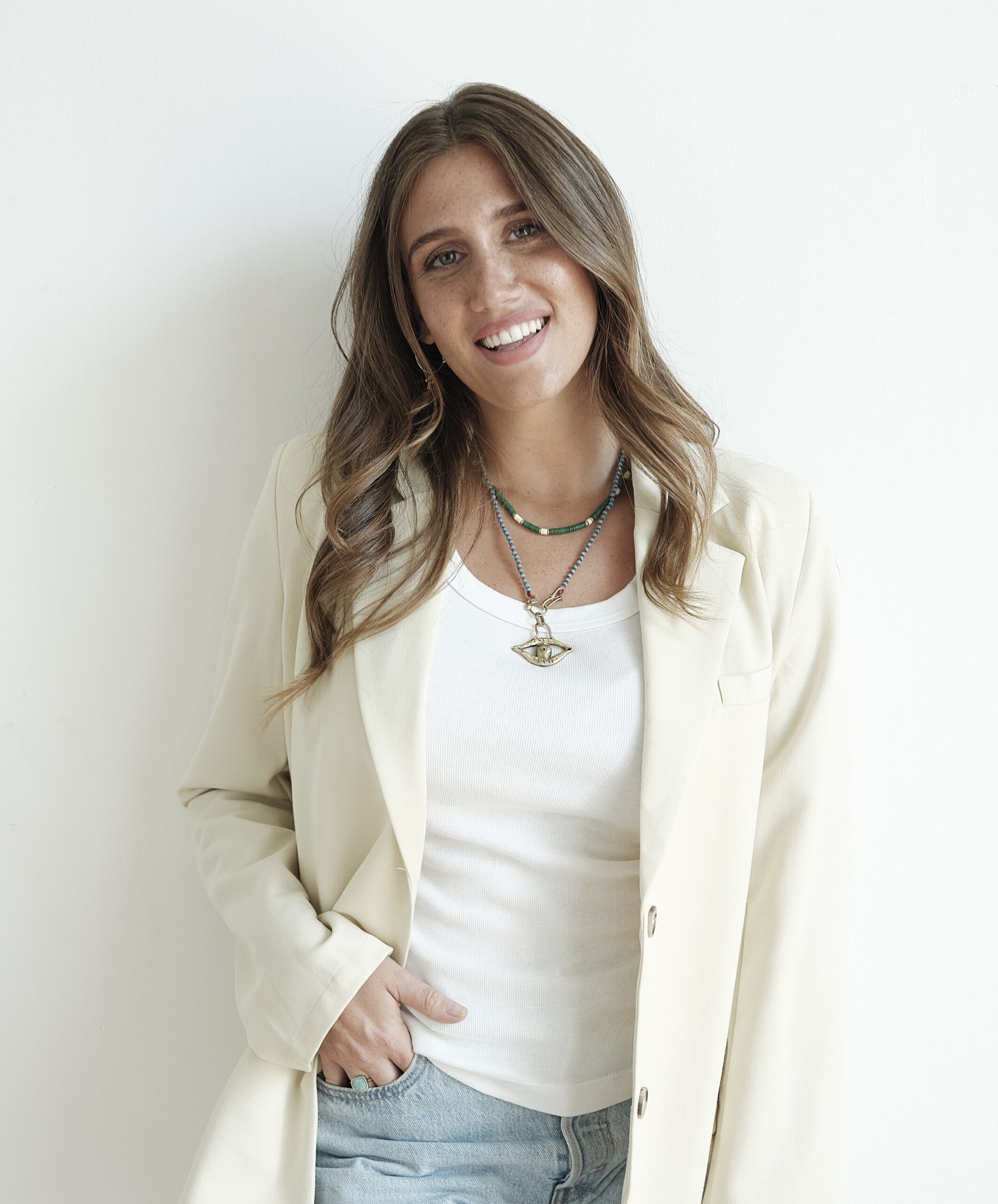 Smiling woman in white blazer and casual outfit posing against a white wall.
