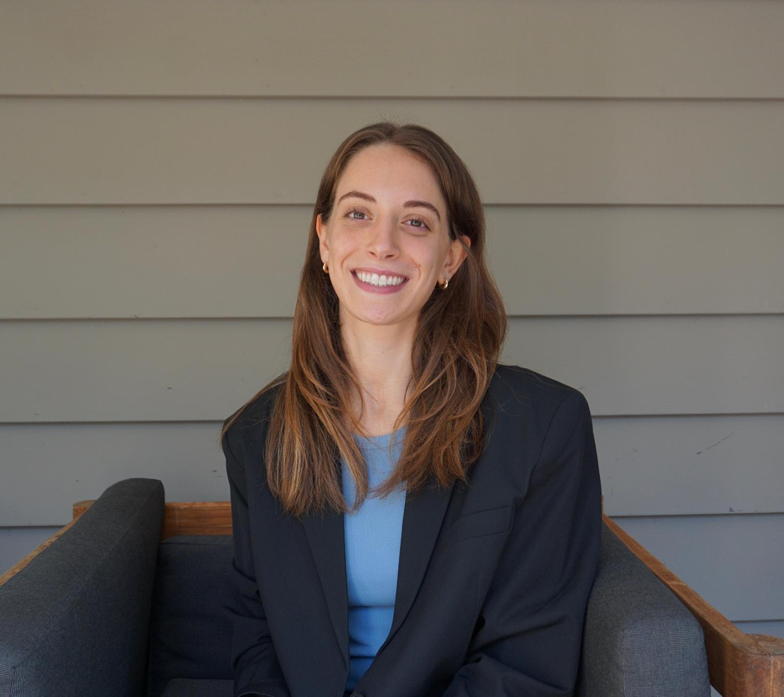 Smiling woman in a blazer sitting on a couch against a paneled wall.