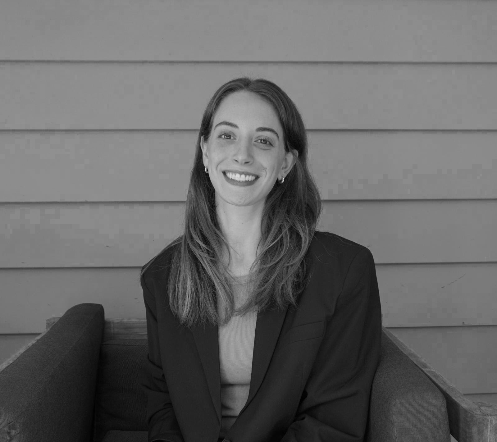 Smiling woman in a blazer sitting on a couch against a paneled wall.