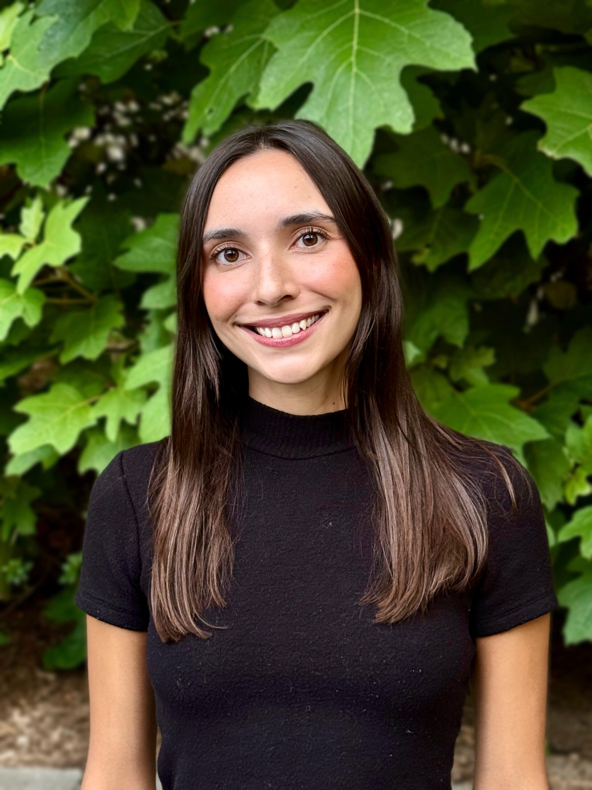 A smiling woman with long brown hair wearing a black shirt stands against a leafy green background.