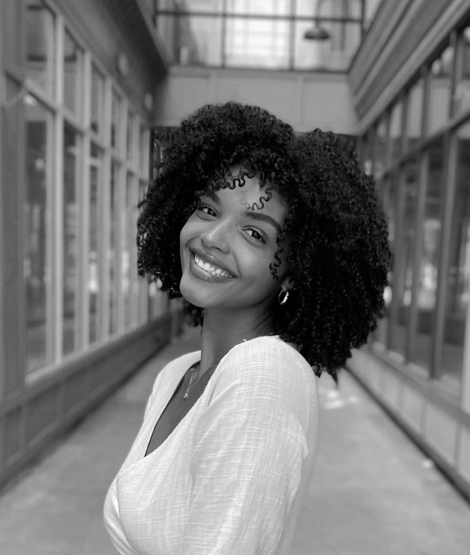 Smiling woman with curly hair in a bright corridor.